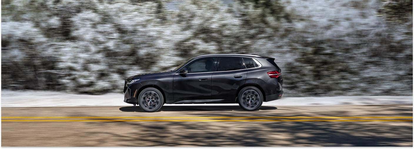 Dark gray 2026 BMW X3 SUV driving along a road in side profile view.