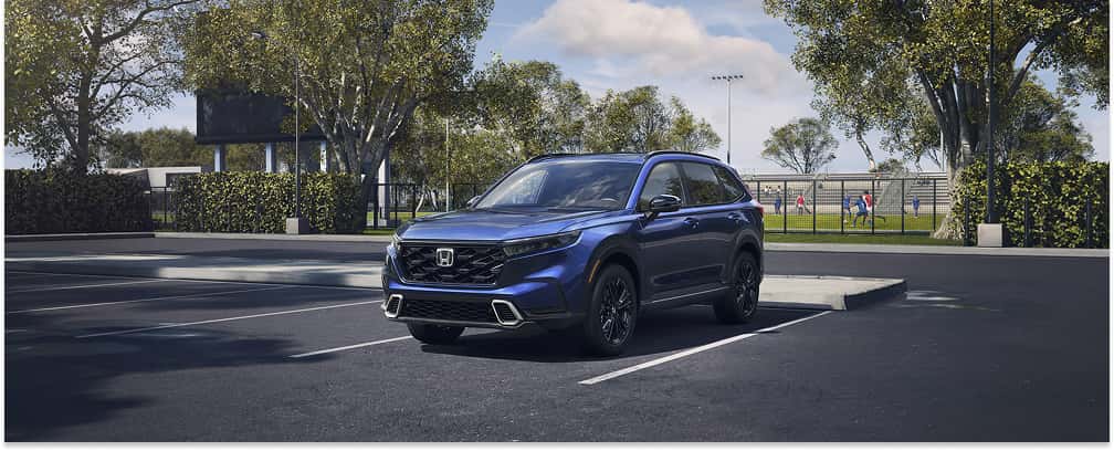 A blue 2026 Honda CR-V SUV is parked in an asphalt parking lot next to an outdoor sports field, set against a background of green trees.