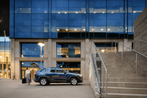 A dark blue Toyota RAV4 parked in front of a modern building with lots of full-length windows while a woman in a business suite closes the trunk.