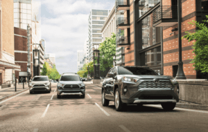 Three new Toyota RAV4's driving down a street with buildings lining each side of them.