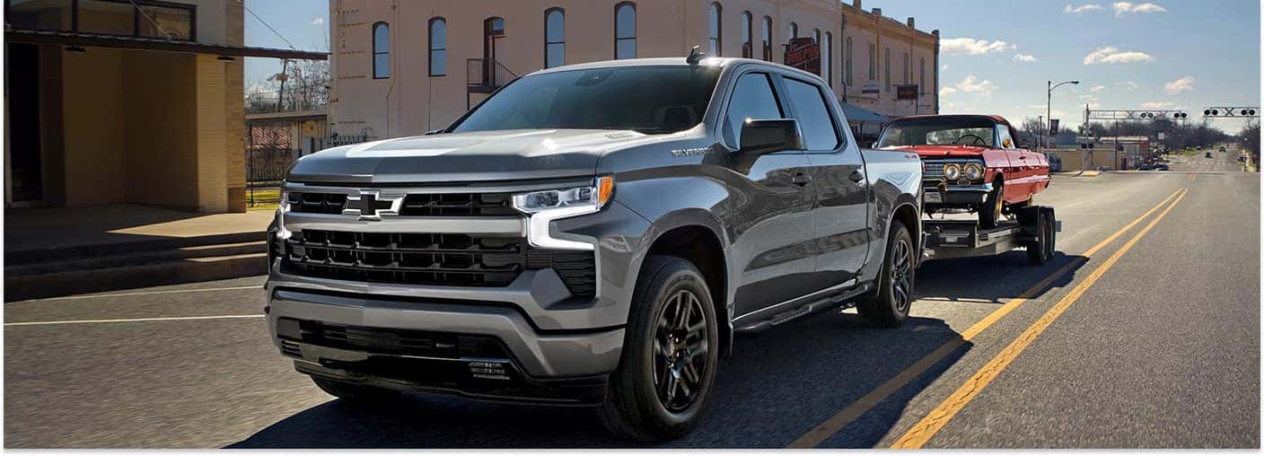 A front three-quarter view of a grey 2026 Chevrolet Silverado 1500 pickup truck towing a classic red car on a flatbed trailer down a paved town street.