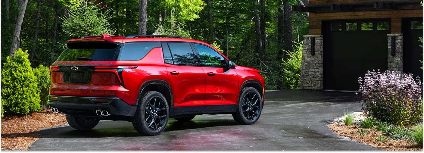 A rear three-quarter view of a red 2026 Chevrolet Traverse parked on a paved driveway in front of a house garage, surrounded by lush green trees and landscaping.