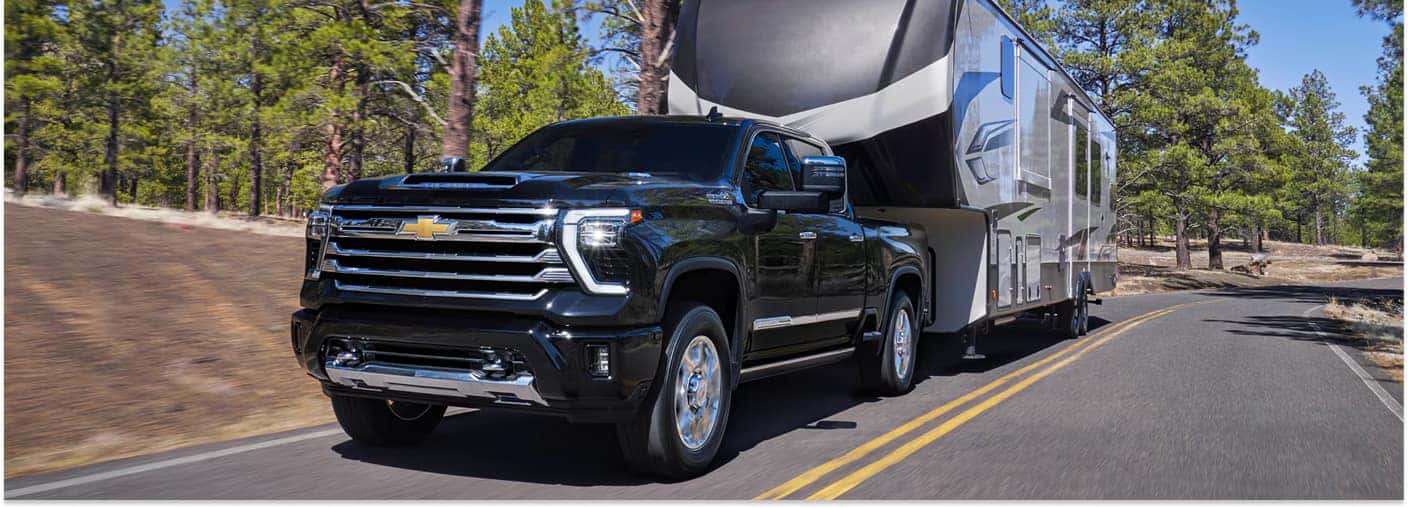Front three-quarter view of a black 2026 Chevrolet Silverado 2500 towing a fifth-wheel trailer along a forest road.
