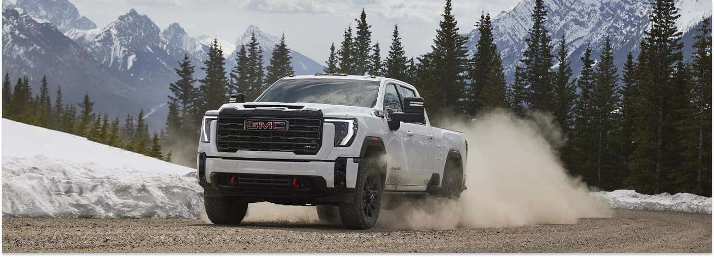 A white 2026 GMC Sierra 2500 heavy-duty pickup truck kicks up a dramatic cloud of dust while driving on a dirt road, set against a majestic backdrop of snow-capped mountains.