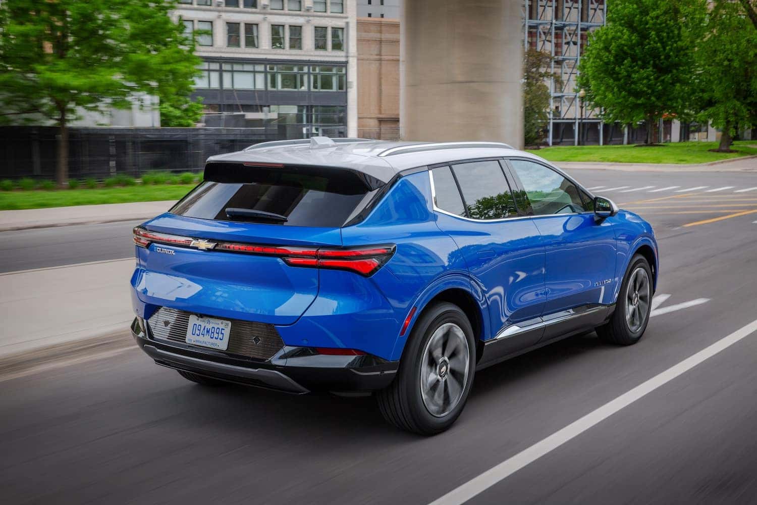 Rear 3/4 view of a 2024 Chevrolet Equinox EV LT in Riptide Blue Metallic with a Summit White roof driving in a downtown area.