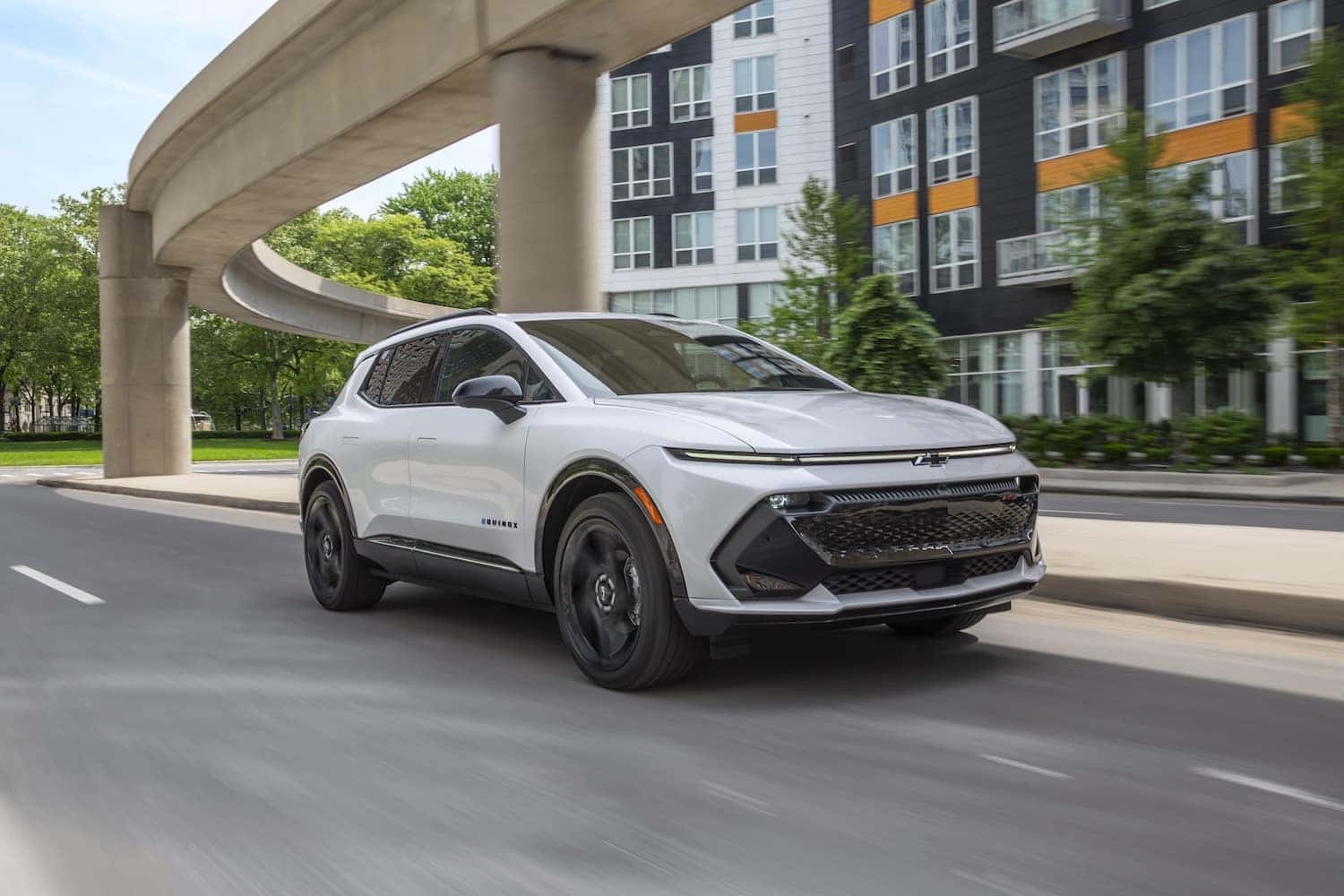 Front 3/4 view of a 2024 Chevrolet Equinox EV RS in Summit White driving in a downtown area.