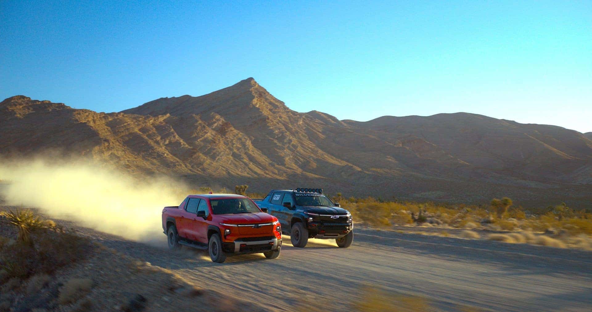Two 2025 Chevrolet Silverado EV ZR2 Racing Trucks - Black and Blue, and Habanero Orange 2025 Chevrolet Silverado EV ZR2 Racing Trucks Driving Side by Side Down the Road in the Mountains