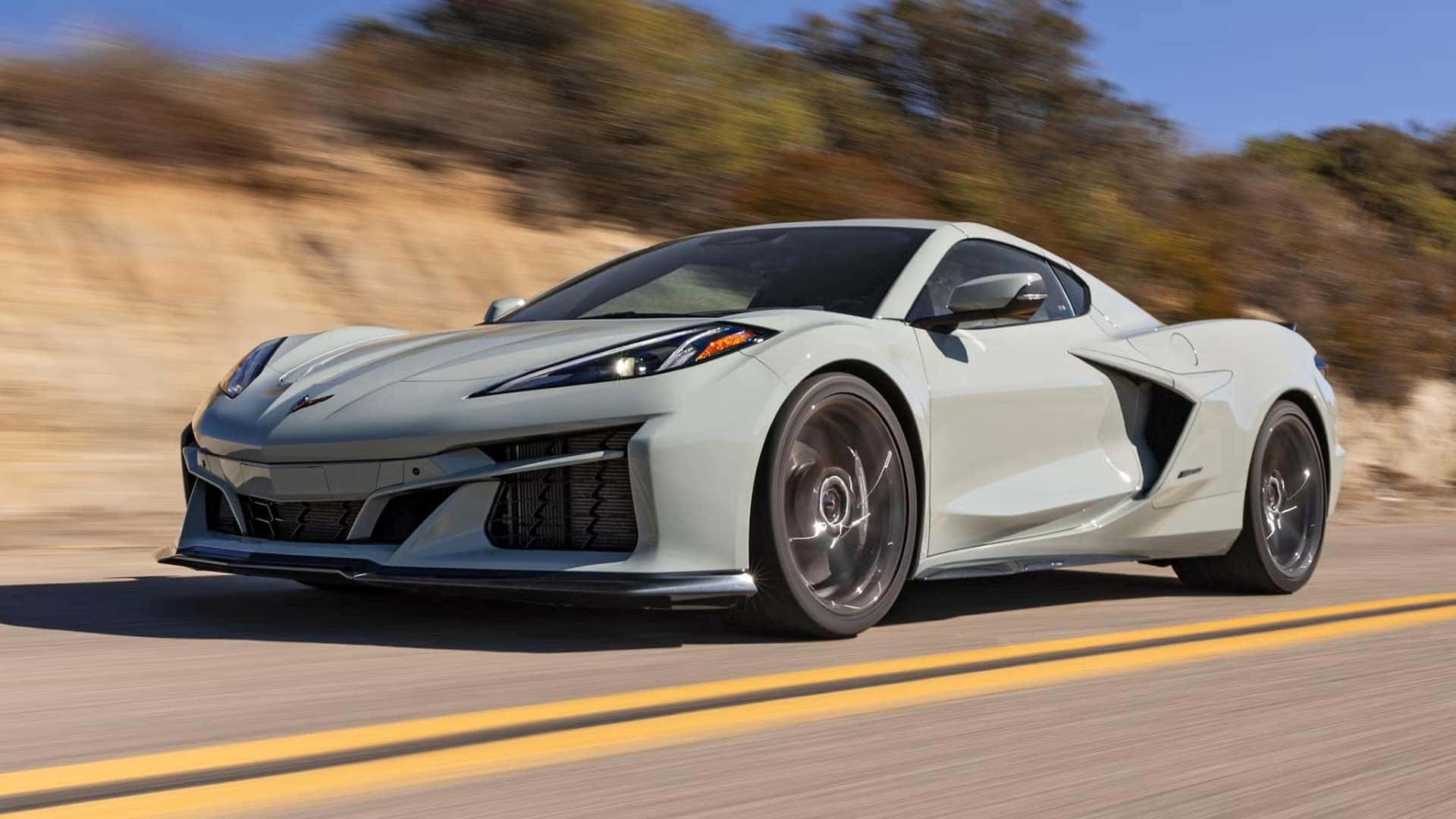 The Gray And Sleek Exterior Chevrolet Corvette E-Ray Driving On The Road Past Rising Hills With Bushes