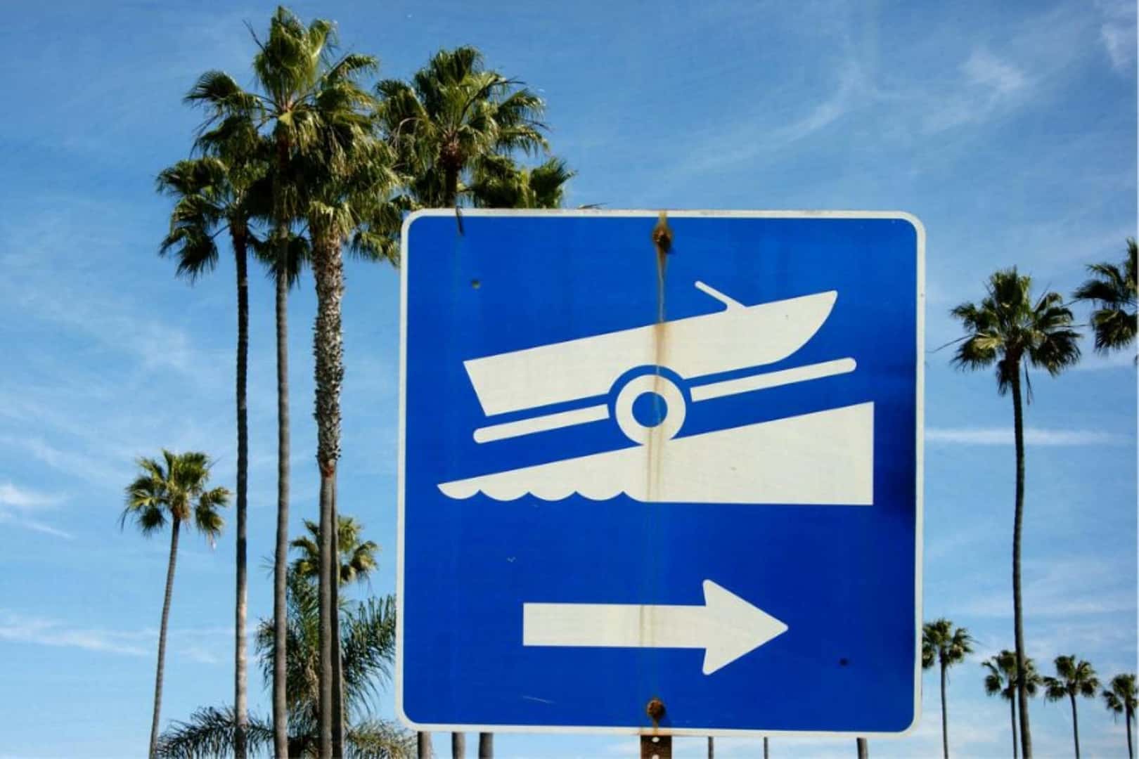 A Blue And White Sign Depicting A Boat Trailer Going Down A Ramp, At A Beach With Palms