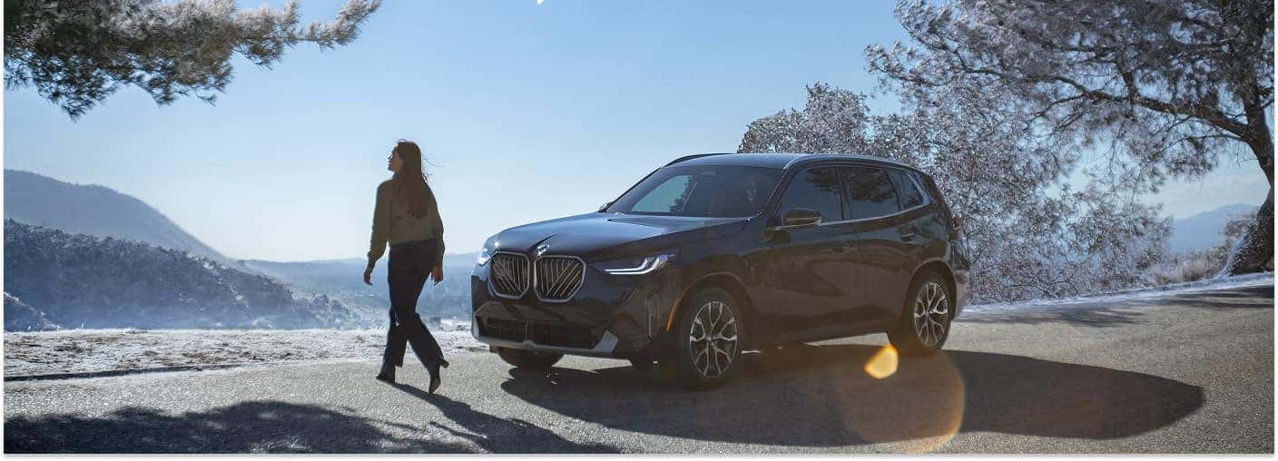 Front three-quarter view of a dark gray 2026 BMW X3 parked on a scenic mountain road.